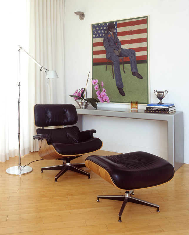 A black leather and plywood lounge chair with its ottoman at a grey console, a framed painting above