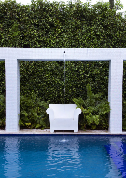 One bay of the pergola, a white armchair centred under a falling water jet against the clipped ficus hedge