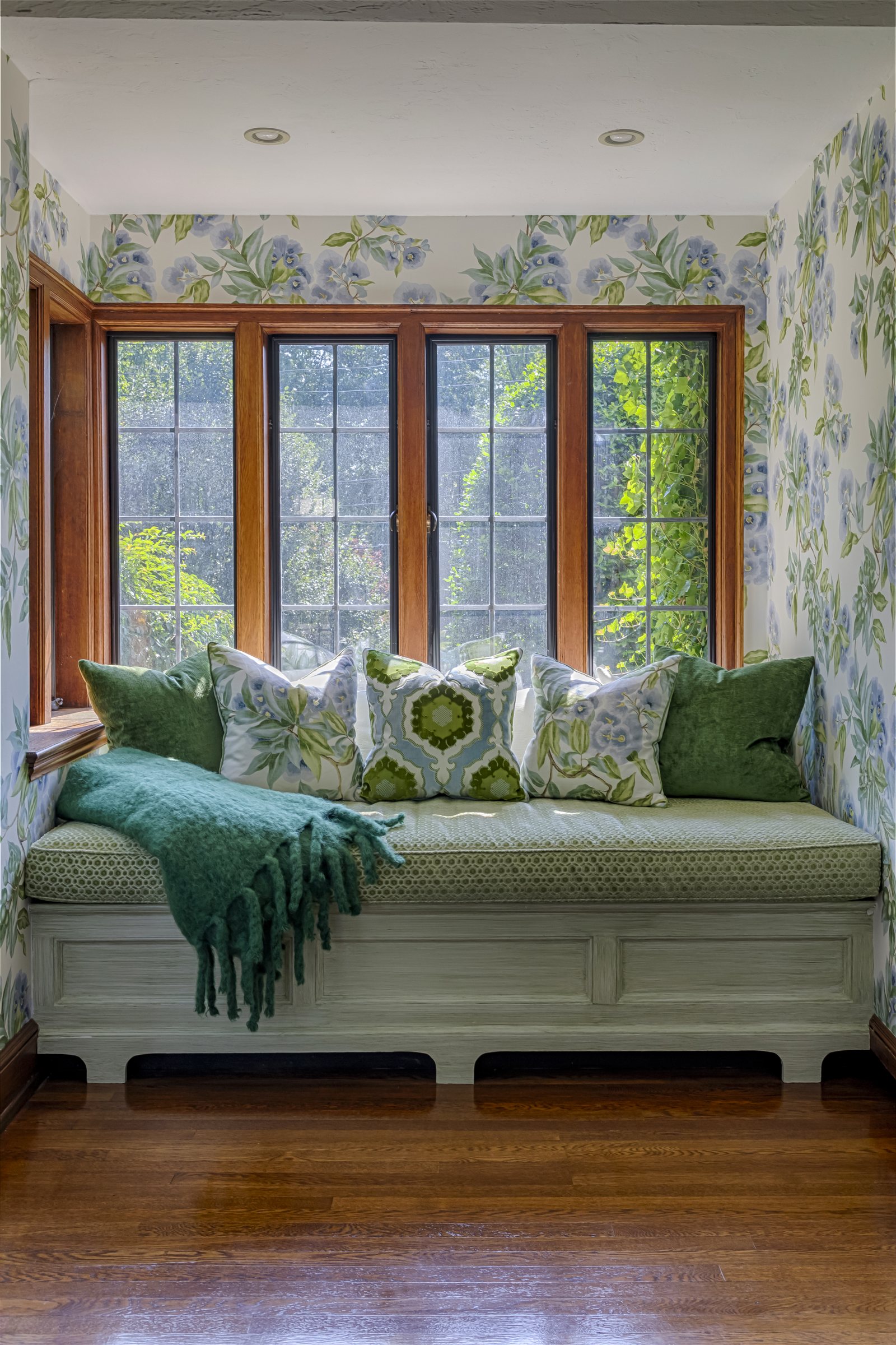 Window seat in an alcove papered in blue and green florals, piled with green and white cushions beneath a bank of wood casements