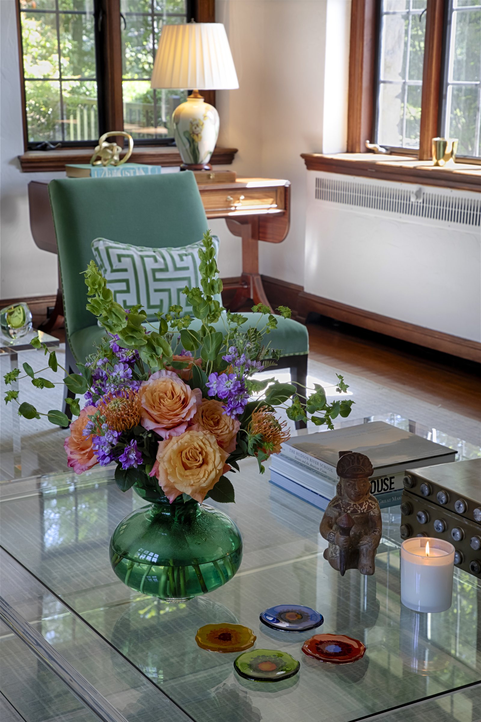 A green glass vase of peach roses and lilac stock on the lucite cocktail table, an aqua chair behind